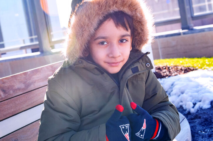 Faiz sits on a wooden bench giving two thumbs up through New England Patriots mittens.