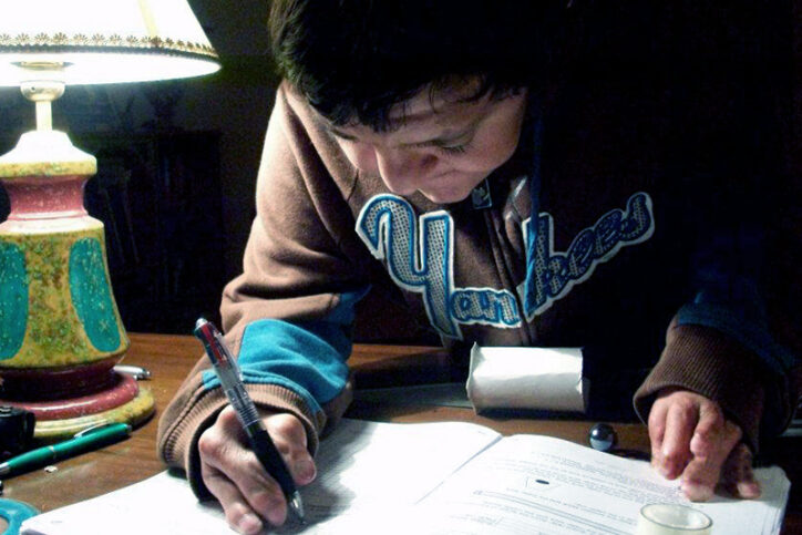 A young man writing in a notebook under a desk lamp.