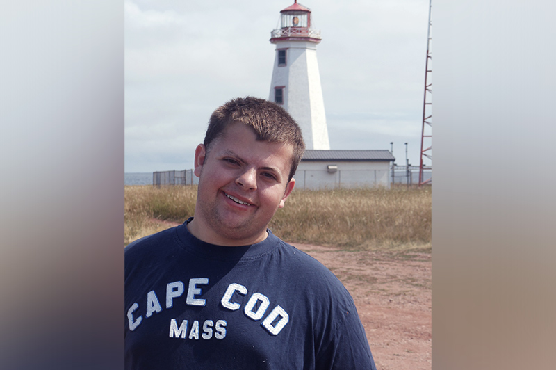 Paul Terry, a patient with Kleefstra syndrome, stands near a lighthouse in a “Cape Cod, Mass” T-shirt.