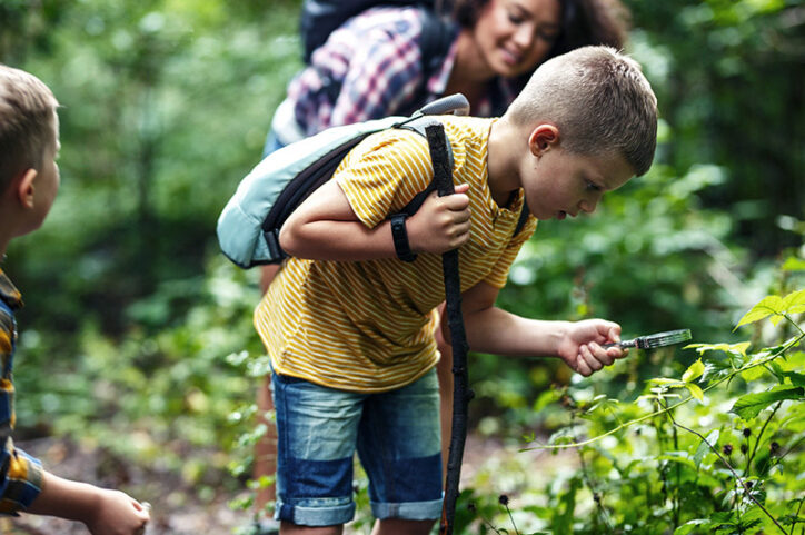 A boy bends over plants in the woods, holding a magnifying glass in his left hand.