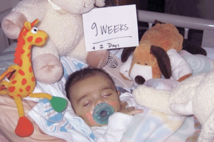 Baby Ben is swaddled in a blanket in a crib, surrounded by stuffed animals. A sign reads "9 weeks old and 2 days".
