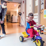 Easton Schlein, dressed in a Spiderman outfit, rides a tricycle in a Boston Children's Hospital hallway.