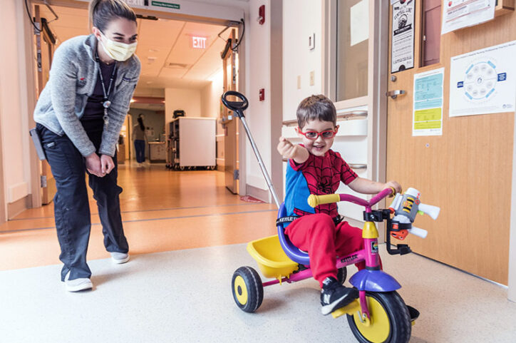 Easton Schlein, dressed in a Spiderman outfit, rides a tricycle in a Boston Children's Hospital hallway.