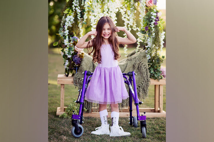 Ashlyn, who has cerebral palsy, in a dress with a matching walker, flexes her arms and smiles.