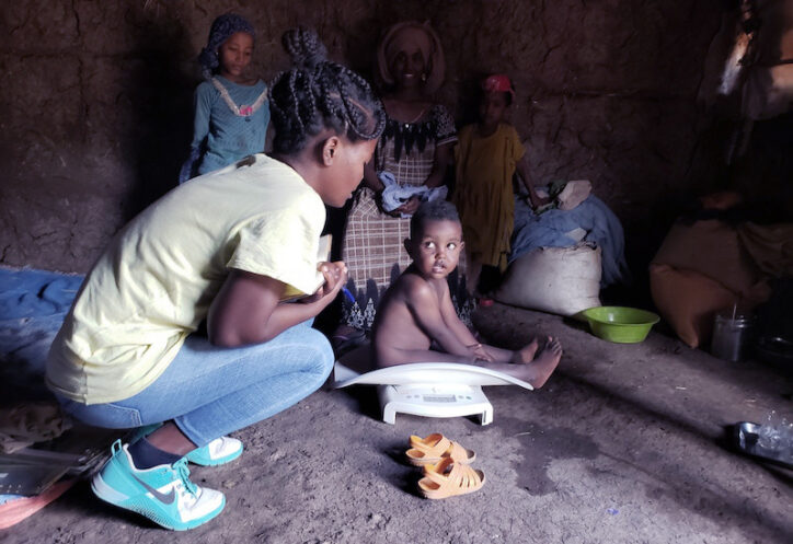 A toddler being weighed on scale on a dirt floor, looking at his mother.
