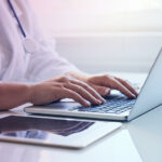 A doctor seated at a desk is typing on a laptop.