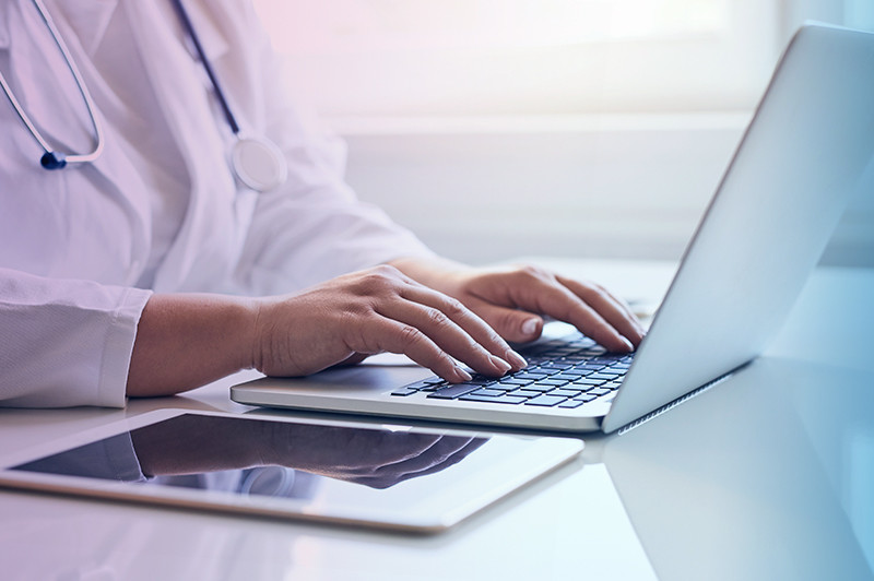 A doctor seated at a desk is typing on a laptop.