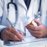 A doctor wearing a white coat is sitting at a desk writing a prescription with a prescription bottle in the other hand.