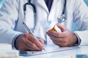 A doctor wearing a white coat is sitting at a desk writing a prescription with a prescription bottle in the other hand.