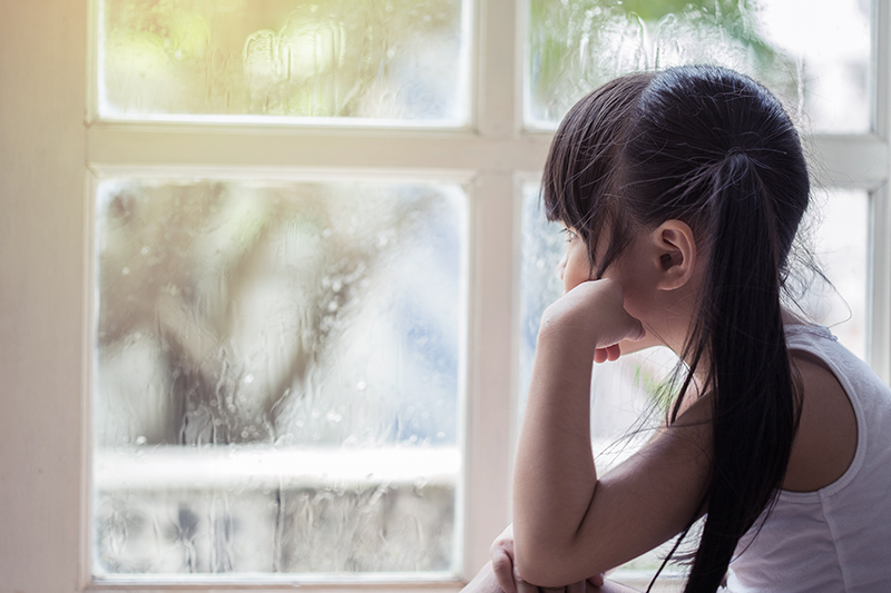 A sad-looking girl at a window on a rainy day.