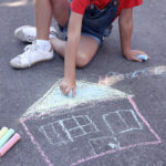 A child draws a chalk illustration of a house on a driveway.