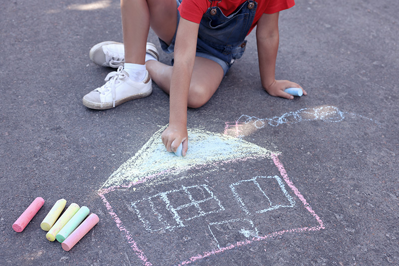 A child draws a chalk illustration of a house on a driveway.