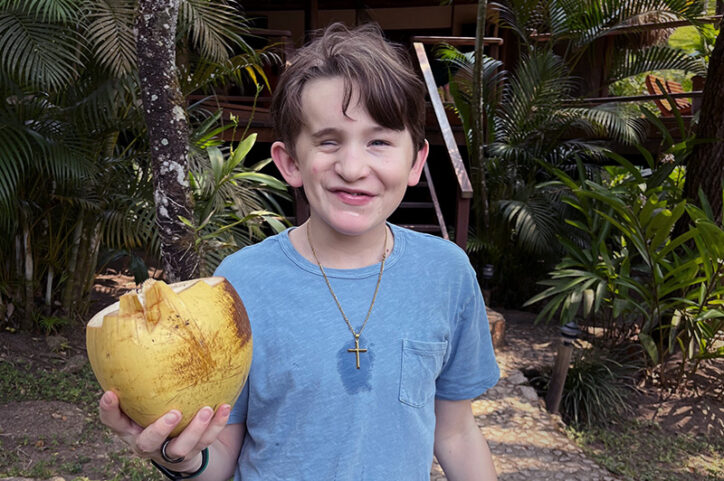 Cruise in front of palm trees holding a coconut and smiling.