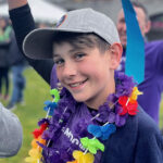 Connor smiles at the camera wearing a rainbow flower necklace, and waves his hands above his head.