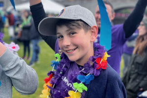 Connor smiles at the camera wearing a rainbow flower necklace, and waves his hands above his head.