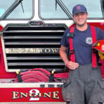 A young man smiling in front of a fire engine holding a firefighter's helmet.