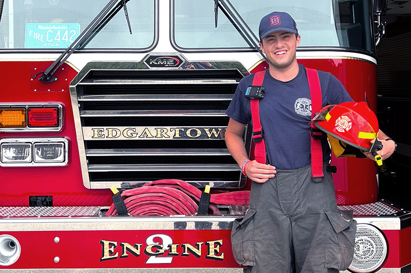 A young man smiling in front of a fire engine holding a firefighter's helmet.