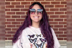 Dianna stands in front of a brick wall smiling. She is wearing sunglasses.