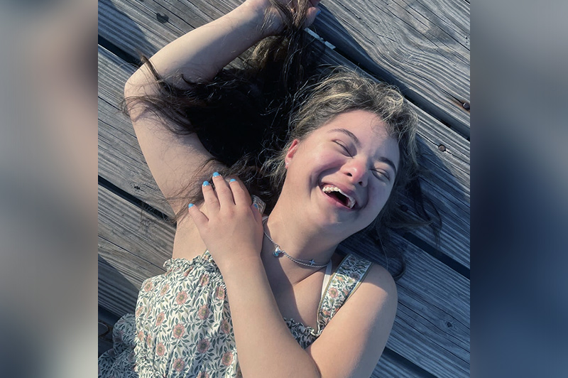 A teenage girl with Down syndrome lies on a porch laughing with her eyes closed.