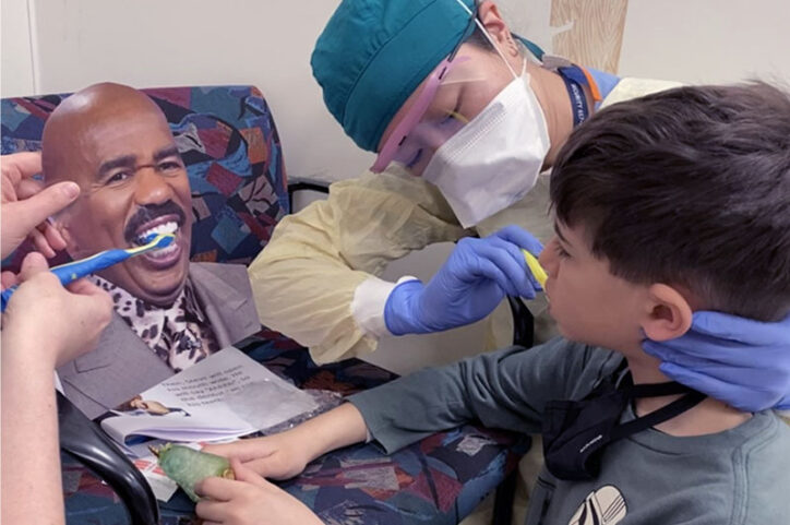 A dentist cleans Panos' teeth while another dentist brushes the teeth of a photo of Steve Harvey.