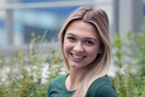 Reagan is smiling at the camera. She's wearing a green shirt, has blonde hair, and is standing in front of green plants.