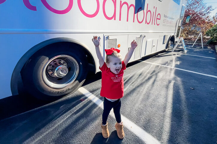 A young girl raises her arm in celebration, standing in front of a blood donation vehicle.