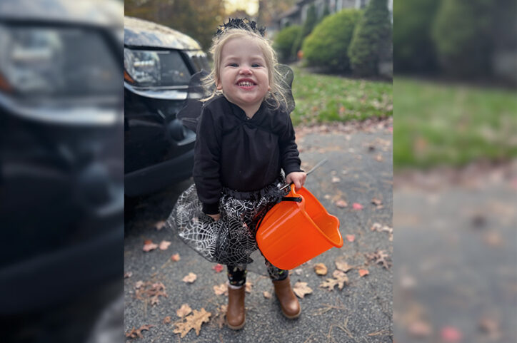 A young girl smiles in her driveway, dressed in a costume and holding a bucket for Halloween candy.