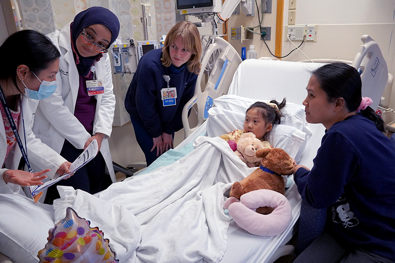 Clinicians gather at the bedside of a young patient, illustrating the concept of family-centered rounds.