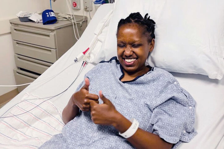 A woman in a hospital bed smiles and gives two-thumbs up.