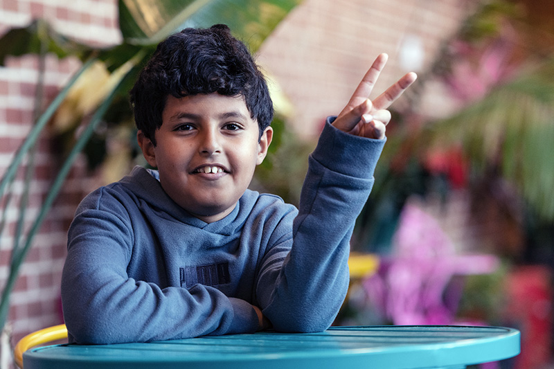 A young boy sitting at a table with two fingers in the air.