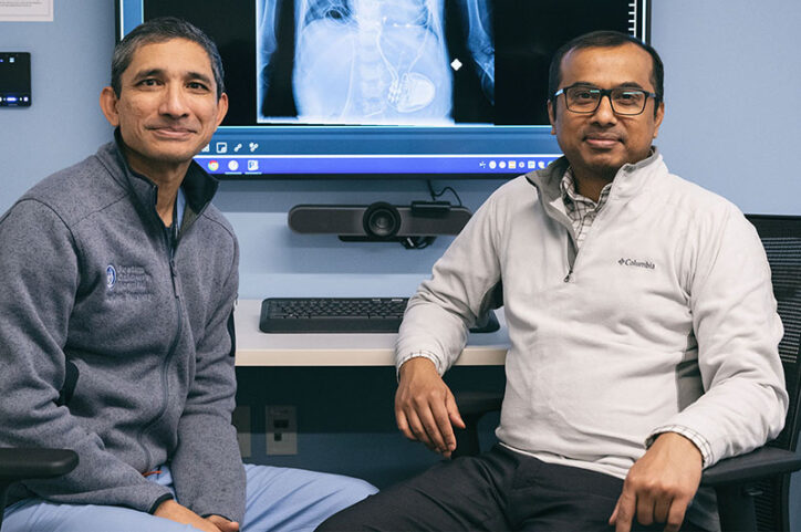 Two doctors sit in front of a large wall-mounted computer screen that is displaying the image of an X-ray.