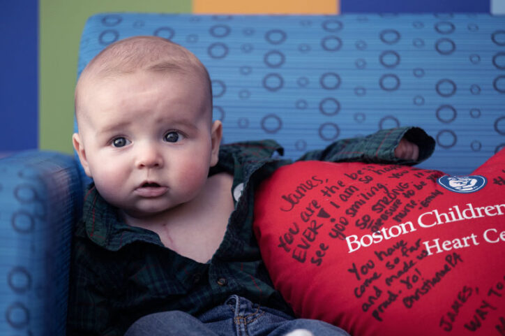 A baby sitting on a couch places his arm on an oversized heart-shaped red pillow.
