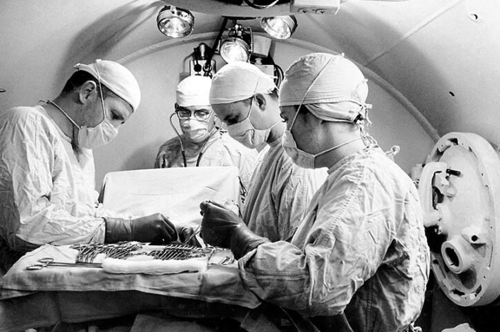 Four medical specialists hover over a tray of medical instruments inside a hyperbaric chamber.