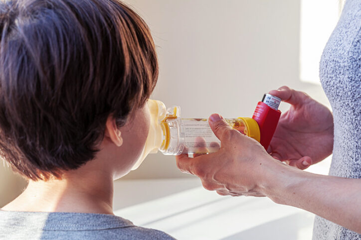 A woman holds a spacer attached to an asthma inhaler while a child takes in the medication.
