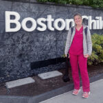 Maeve, who was treated for laryngeal cleft as a child, stands in her nurse scrubs in front of Boston Children's.