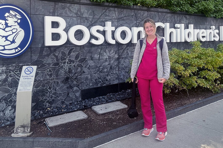 Maeve, who was treated for laryngeal cleft as a child, stands in her nurse scrubs in front of Boston Children's.
