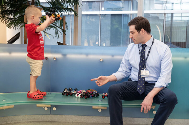 Jack holds aloft a toy truck while standing on a bench. Sitting on the bench is Dr. Matan Setton.