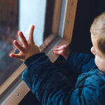 A lonely-appearing toddler looking out a window with his hand pressed against the glass.