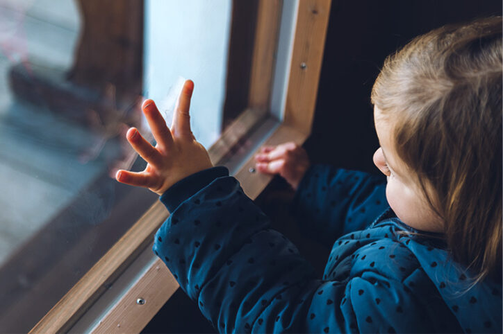 A lonely-appearing toddler looking out a window with his hand pressed against the glass.