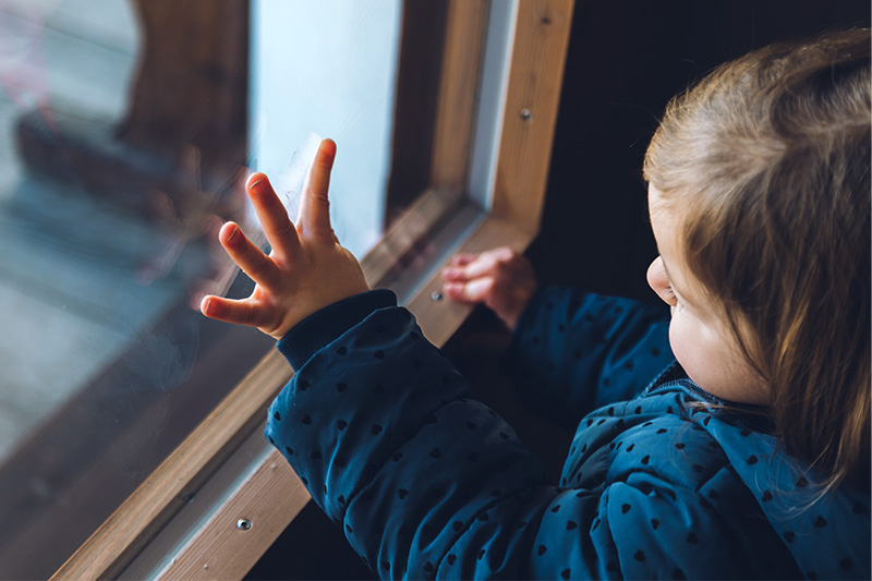 A lonely-appearing toddler looking out a window with his hand pressed against the glass.