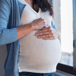 A pregnant women holding her belly by a window.