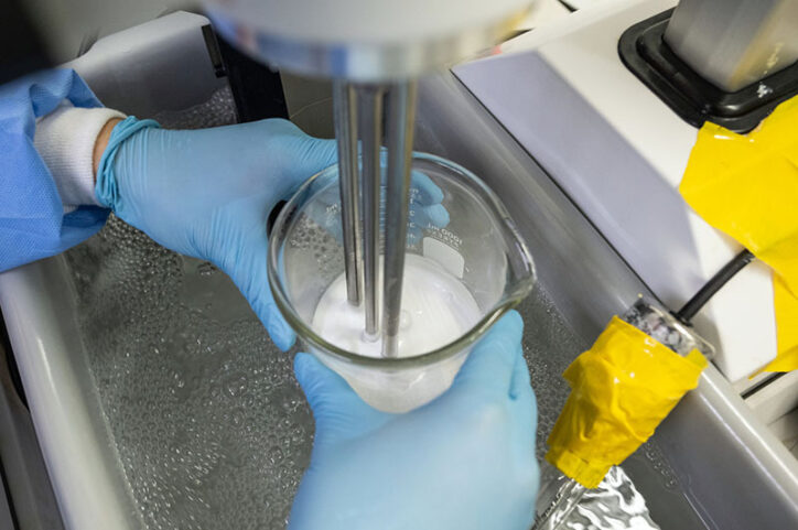 A pair of gloved hands hold a glass container, above which a machine mixes the solution to create gas microbubbles.