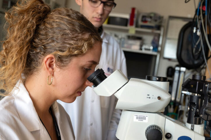 A research assistant looks into a microscope while a colleague stands beside her observing her work.