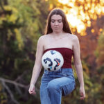 Sadie, a young woman, knee pops a soccer ball. Her hair is down, and her eyes are focused on the ball. She is outdoors.