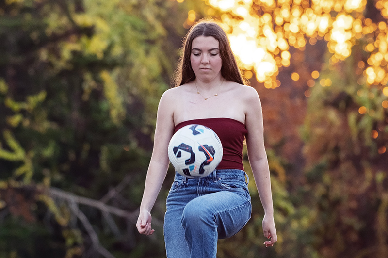 Sadie, a young woman, knee pops a soccer ball. Her hair is down, and her eyes are focused on the ball. She is outdoors.