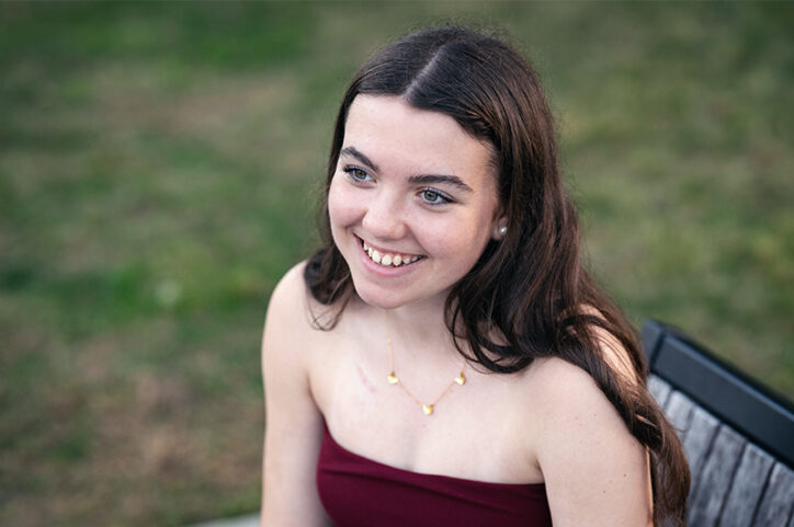 Sadie is sitting in a chair outdoors. Her hair is down. She is looking past the camera and smiling confidently. 