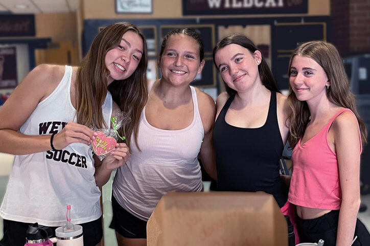 Four young women stand side by side with their arms around each other's backs, smiling.