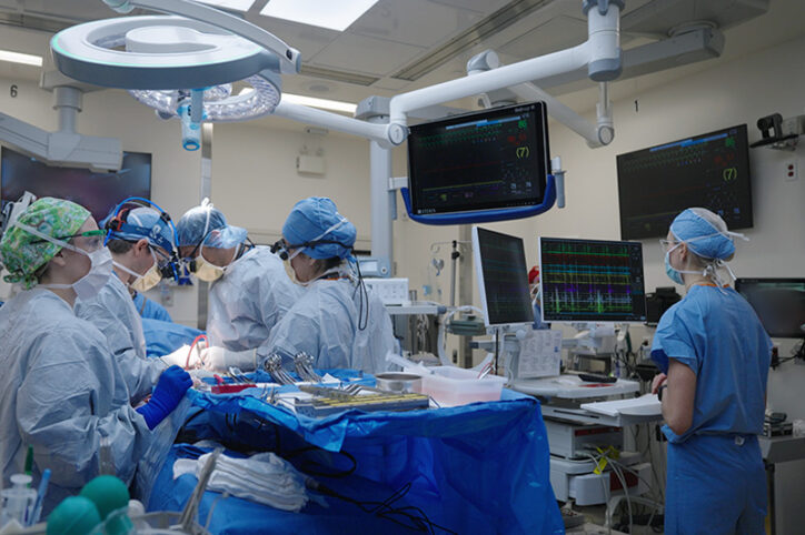 Heart surgeons operate on a patient, while to their right, a cardiologist reads a monitor to reduce heart block.