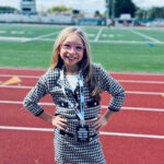 A smiling 10-year-old girl stands, hands on her hips, on the sidelines of a football field.