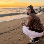 A young woman squats on the shoreline of a beach at sunset.